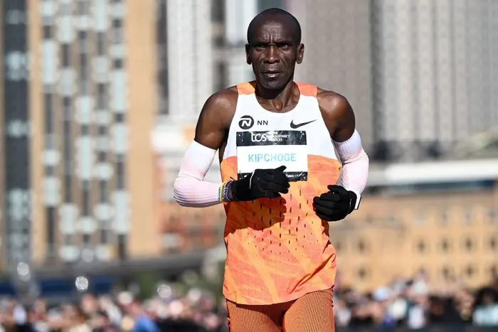 Eliud Kipchoge corriendo un maratón con equipación naranja y mangas blancas, enfocado y concentrado.