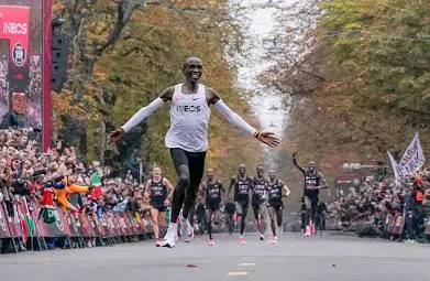 Eliud Kipchoge cruzando la línea de meta sonriendo con los brazos abiertos en el reto INEOS 1:59 en Viena, rodeado de público y liebres de carrera.